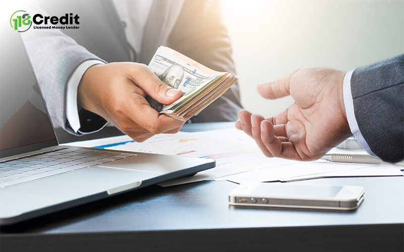 Person handing over a stack of $100 bills across a desk with a laptop, documents, and smartphone.
