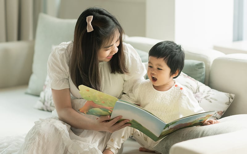 Mother reading a storybook to her toddler on the sofa.